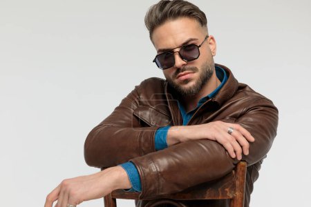 Foto de Portrait of a handsome casual man resting his arms and looking at the camera, sitting on a chair, wearing sunglasses against gray studio background - Imagen libre de derechos