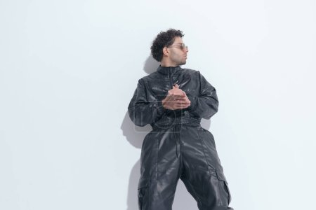 Téléchargez les photos : Sexy man with curly hair in leather suit rubbing palms and looking to side on grey background in studio - en image libre de droit