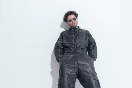 Photo for Greek fashion model with curly hair in leather suit and sunglasses holding hands in pockets, laying on a wall and posing in front of grey background - Royalty Free Image