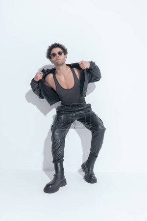 Photo for Curly young arab man with sunglasses adjusting and pulling leather jacket while laying on a wall on grey background - Royalty Free Image
