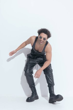 Photo for Happy muscle man with curly hair wearing undershirt and leather pants, laying on a wall and laughing on grey background - Royalty Free Image