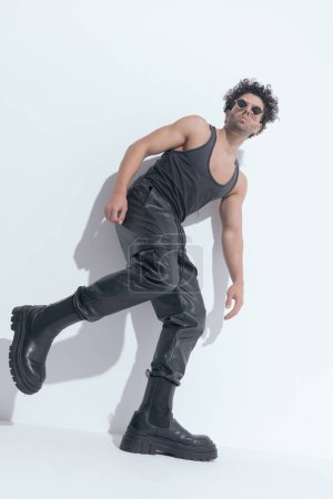 Foto de Hombre fresco en forma usando pantalones de cuero y camiseta, acostado en una pared y sosteniendo una pierna hacia arriba mientras posando en una pose vista lateral - Imagen libre de derechos