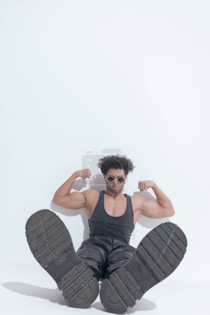 Téléchargez les photos : Puissant jeune homme flexion biceps et montrant des bottes tout en étant assis sur fond gris - en image libre de droit