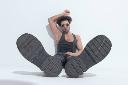 Téléchargez les photos : Homme de mode en maillot de corps tenant le bras au-dessus de la tête, assis et montrant des bottes sur fond gris - en image libre de droit