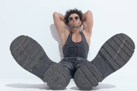 Téléchargez les photos : Fit young man with curly hair in undershirt sitting and showing boots while posing with hands behind neck - en image libre de droit