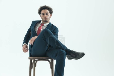Foto de Retrato de un hombre de negocios guapo cruzando las piernas y sintiéndose relajado, sentado, usando anteojos contra el fondo gris del estudio - Imagen libre de derechos