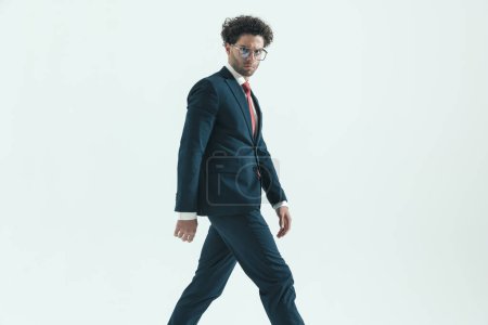 Photo for Side view of sexy businessman walking one way while looking at camera wearing eyeglasses against gray studio background - Royalty Free Image