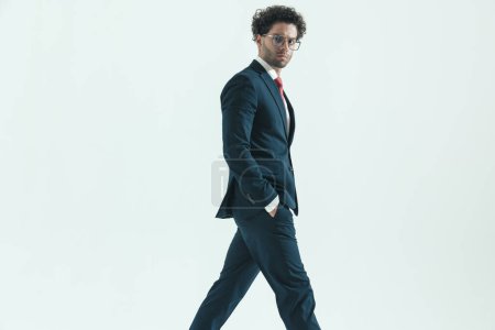 Photo for Side view of attractive businessman walking on his path wih hands in pockets wearing eyeglasses against gray studio background - Royalty Free Image