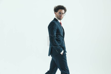 Foto de Vista lateral del joven hombre de negocios caminando con las manos en los bolsillos y mirada dura con gafas graduadas sobre fondo gris estudio - Imagen libre de derechos