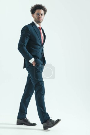 Photo for Full body picture of attractive businessman walking with hands in pockets and tough face wearing eyeglasses against gray studio background - Royalty Free Image