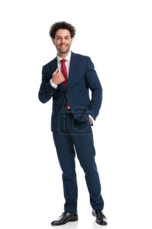 Foto de Happy young man in elegant suit smiling, holding hand in pocket and fixing suit in front of white background in studio - Imagen libre de derechos