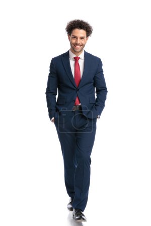 Téléchargez les photos : Élégant jeune homme d'affaires en costume bleu marine marchant les mains dans les poches souriantes et posant sur fond blanc en studio - en image libre de droit