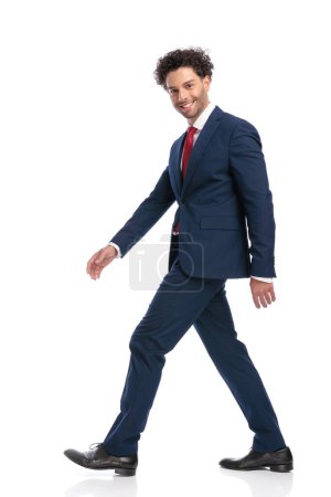 Foto de Vista lateral de hombre de negocios feliz en traje sonriendo y caminando delante de fondo blanco en el estudio - Imagen libre de derechos