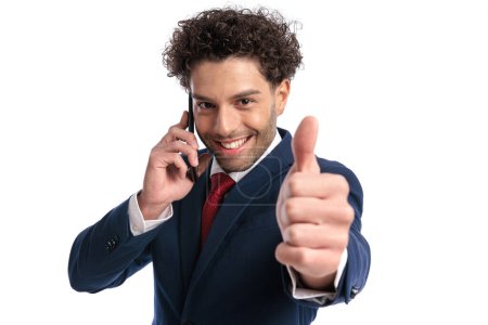 Photo for Curly hair man having a mobile conversation and making thumbs up sign in front of white background in studio - Royalty Free Image