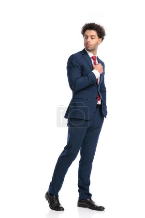 Foto de Elegante joven hombre de negocios mirando por encima del hombro y arreglando corbata delante de fondo blanco - Imagen libre de derechos