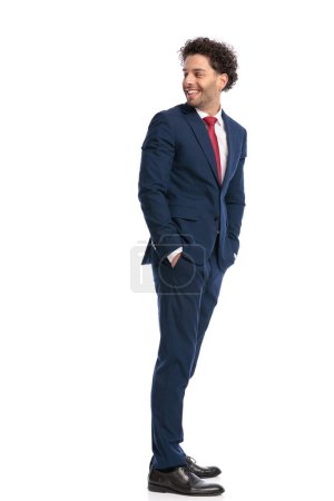 Foto de Feliz hombre de negocios elegante cogido de la mano en los bolsillos y mirando por encima del hombro y sonriendo delante de fondo blanco en el estudio - Imagen libre de derechos