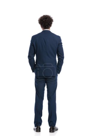 Photo for Rear view of elegant man in navy blue suit holding hands in pockets in front of white background in studio - Royalty Free Image