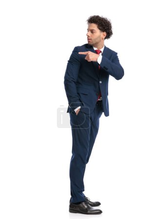 Foto de Side view of elegant curly hair man looking behind and pointing finger while standing and posing on white background in studio - Imagen libre de derechos