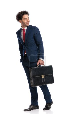 Foto de Hombre guapo con maleta mirando por encima del hombro y riendo delante de fondo blanco en el estudio - Imagen libre de derechos