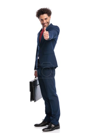 Téléchargez les photos : Happy elegant businessman smiling, holding suitcase and making thumbs up gesture on white background in studio - en image libre de droit