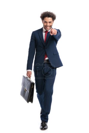 Téléchargez les photos : Jeune homme d'affaires libanais tenant une valise, pointant du doigt et riant tout en marchant sur fond blanc en studio - en image libre de droit