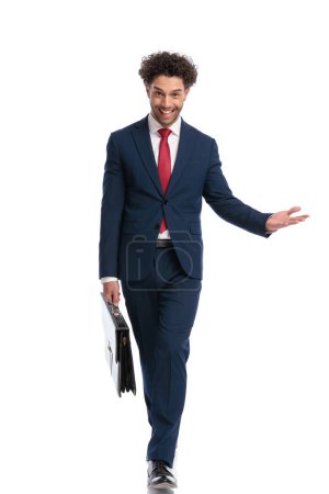 Foto de Happy elegant man wearing suit, holding bag and inviting to side while walking in front of white background in studio - Imagen libre de derechos