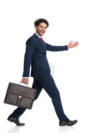 Foto de Elegant arabic man in suit with suitcase walking and welcoming with a happy face in front of white background in studio - Imagen libre de derechos