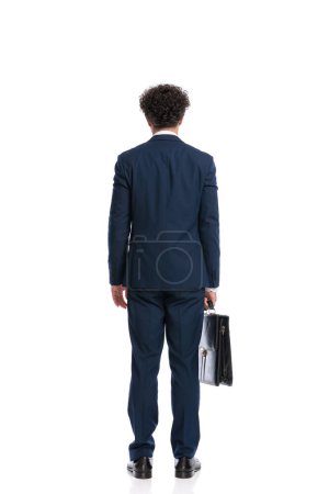 Photo pour Rear view of elegant young businessman in suit holding briefcase in front of white background in studio - image libre de droit