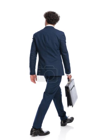 Foto de Back view of elegant businessman in suit holding suitcase and walking in front of white background in studio - Imagen libre de derechos