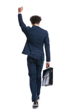 Téléchargez les photos : Derrière la vue du jeune homme d'affaires tenant le poing vers le haut et célébrant tout en tenant la valise et marchant sur fond blanc - en image libre de droit