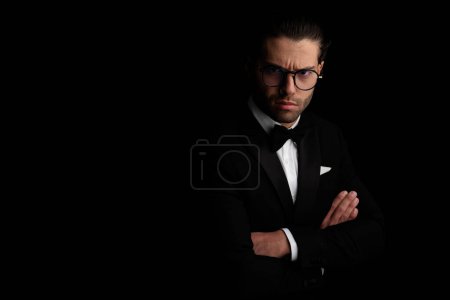 Photo for Portrait of frowning elegant young man in black tuxedo with eyeglasses crossing arms and posing on black background - Royalty Free Image
