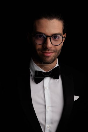 Téléchargez les photos : Bel homme élégant portant smoking noir et lunettes et souriant devant fond noir en studio, portrait - en image libre de droit