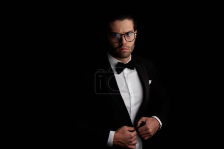 Photo for Sexy young best man with eyeglasses adjusting and closing black tuxedo in front of black background in studio - Royalty Free Image