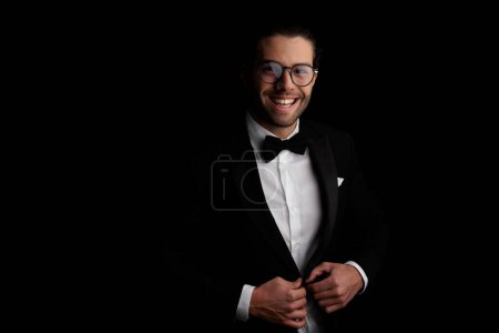 Téléchargez les photos : Portrait de beau jeune marié avec des lunettes déboutonnant smoking et riant devant fond noir en studio - en image libre de droit