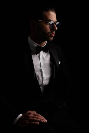 Photo for Portrait of sexy young man in tuxedo looking to side and posing in a mysterious way on black background in studio - Royalty Free Image