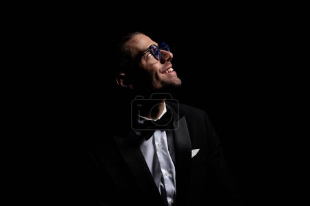 Téléchargez les photos : Heureux marié élégant en smoking noir avec des lunettes levant les yeux, trouver la lumière dans l'obscurité devant fond noir - en image libre de droit