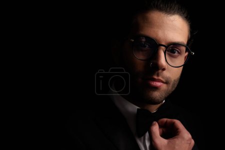Téléchargez les photos : Attrayant jeune homme élégant avec des lunettes ajustant noeud papillon et souriant devant fond noir - en image libre de droit