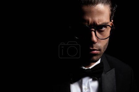 Photo for Close up portrait of handsome turkish man with glassess wearing tuxedo and posing in a mysterious way with light on black background - Royalty Free Image