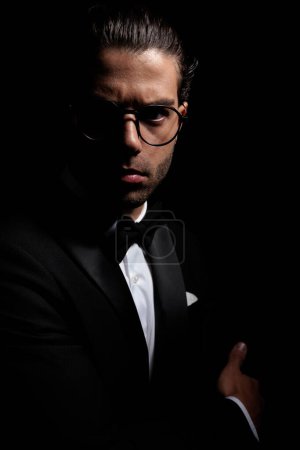 Photo for Elegant young groom with glasses posing in a mysterious light with folded arms on black background - Royalty Free Image