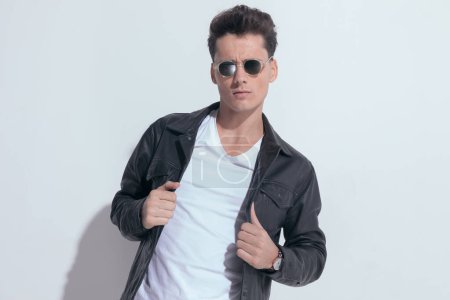 Photo for Portrait of  young casual man pulling his jacket with tough attitude, standing, wearing sunglasses in a fashion pose - Royalty Free Image