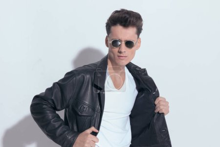 Téléchargez les photos : Portrait of  attractive casual man with macho style is pulling his leather jacket, standing, wearing sunglasses in a fashion pose - en image libre de droit
