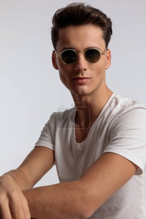 Photo for Portrait of attractive casual man rocking a pair of sunglasses, sitting, wearing sunglasses in a fashion pose - Royalty Free Image