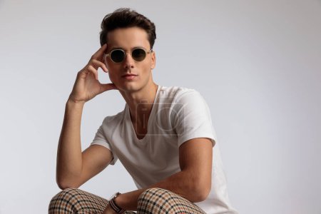 Téléchargez les photos : Portrait d'homme décontracté attrayant touchant son visage et posant avec style, assis, portant des lunettes de soleil dans une pose de mode - en image libre de droit
