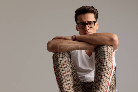 Téléchargez les photos : Portrait de jeune homme décontracté mettant la main sur ses genoux, assis, portant des lunettes dans une pose de mode - en image libre de droit