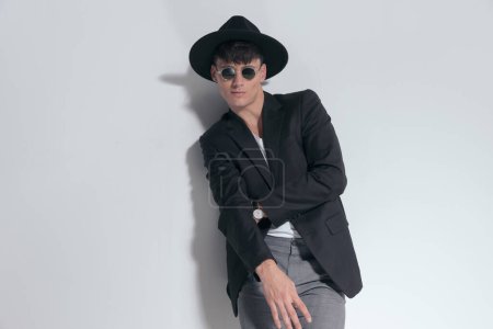 Photo for Portrait of young smart casual man crossing his hands and smiling, standing, wearing a black hat and sunglasses in a fashion pose - Royalty Free Image