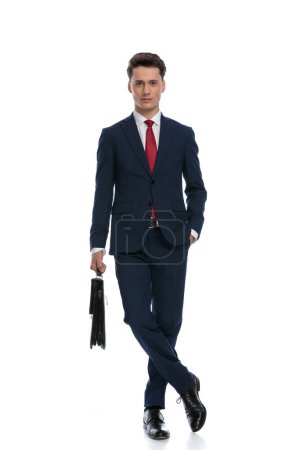 Photo for Handsome businessman crossing legs, holding a briefcase with hand in pocket, wearing a suit and tie against white background - Royalty Free Image