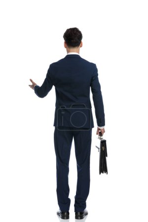 Téléchargez les photos : Élégant homme d'affaires tenant la mallette et présentant, portant un costume et cravate sur fond blanc - en image libre de droit