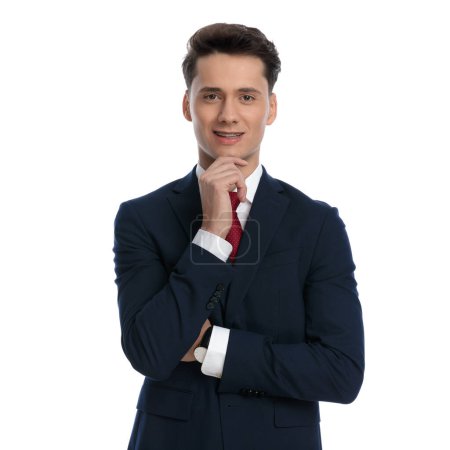 Téléchargez les photos : Jeune homme d'affaires croisant les bras, griffant le menton et souriant, portant un costume et une cravate sur fond blanc - en image libre de droit