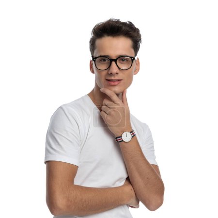Foto de Retrato de hombre guapo con gafas cogidas de la mano a la barbilla y pensando mientras posando frente al fondo blanco en el estudio - Imagen libre de derechos