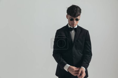 Photo for Portrait of handsome businessman fixing his sleeve and looking down, standing, wearing a black tuxedo and sunglasses, in a fashion pose - Royalty Free Image